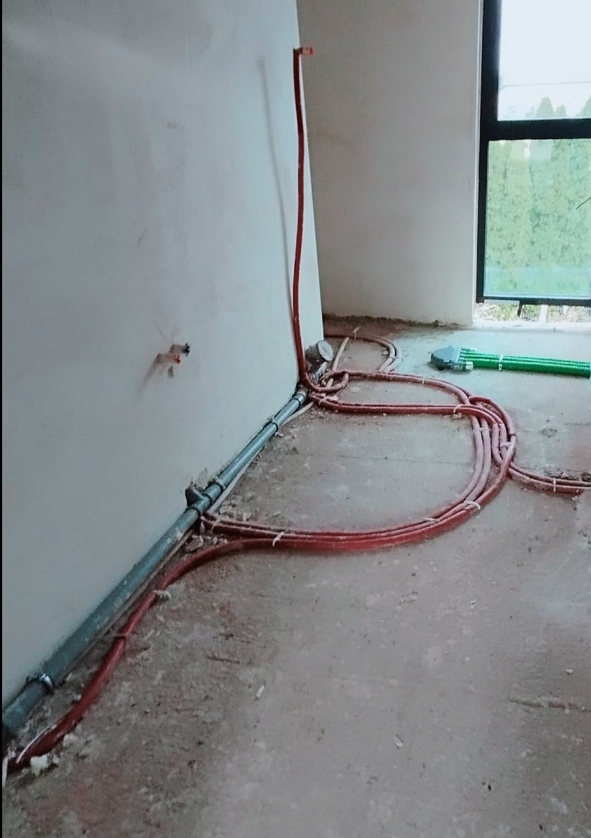 Wnętrze w trakcie montażu instalacji ogrzewania podłogowego: czerwone rury grzewcze ułożone na surowym betonie, szara rura wzdłuż ściany, widok na okno z zielenią na zewnątrz.