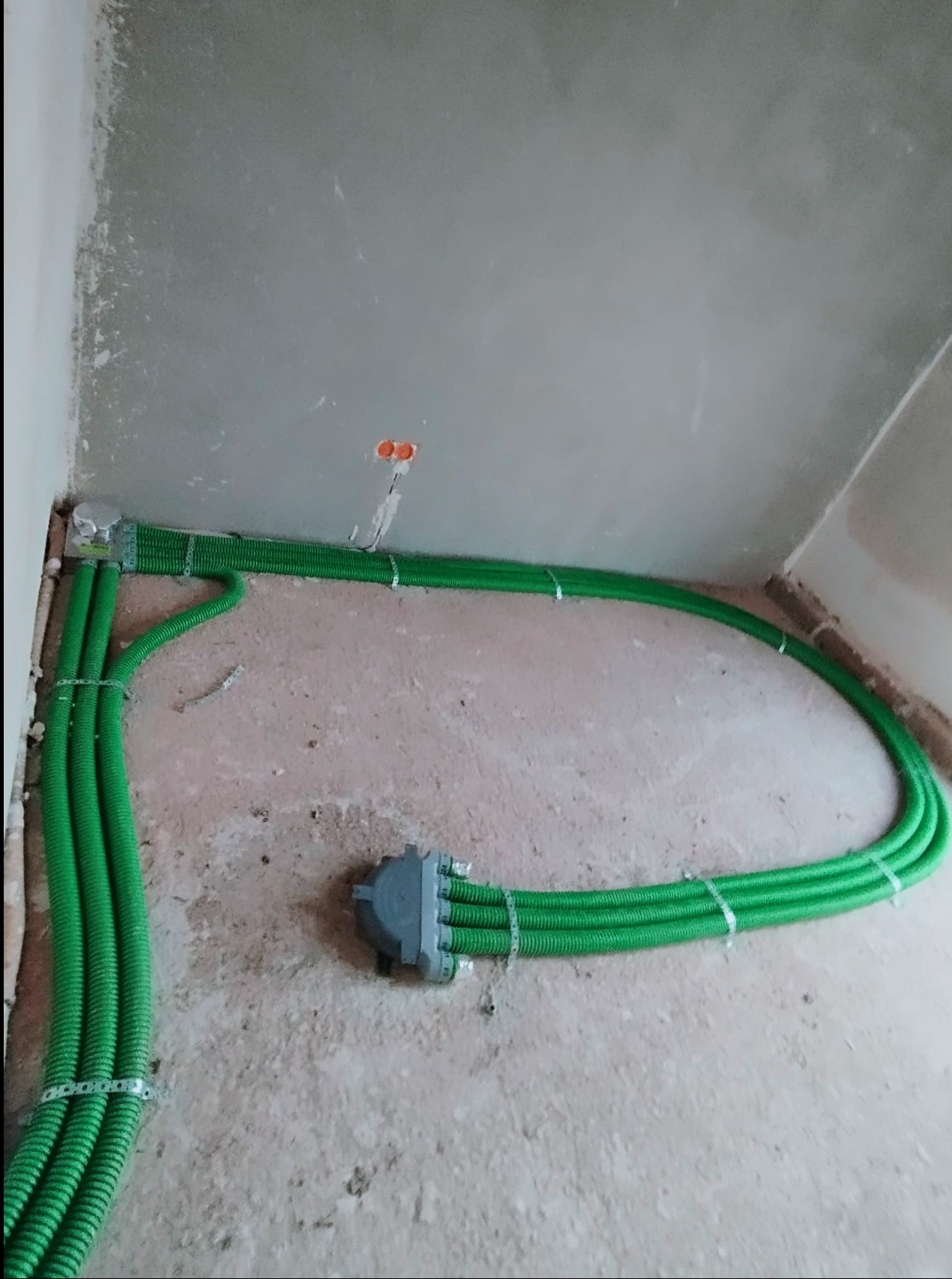 Zielone rury karbowane prowadzone po ścianie i suficie w surowym stanie wykończenia wnętrza, mocowane metalowymi uchwytami. Widoczne puszki elektryczne w ścianie.