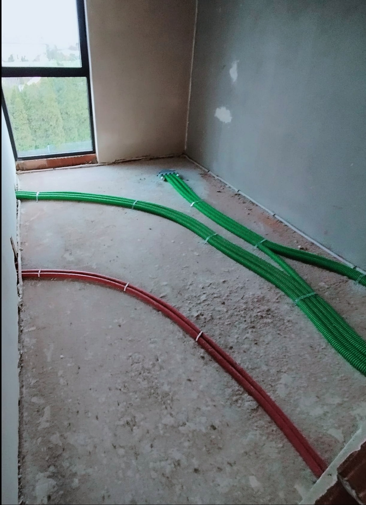 Wnętrze w trakcie prac instalacyjnych w Rybniku: zielone i czerwone rury prowadzone po surowej posadzce w kierunku okna.