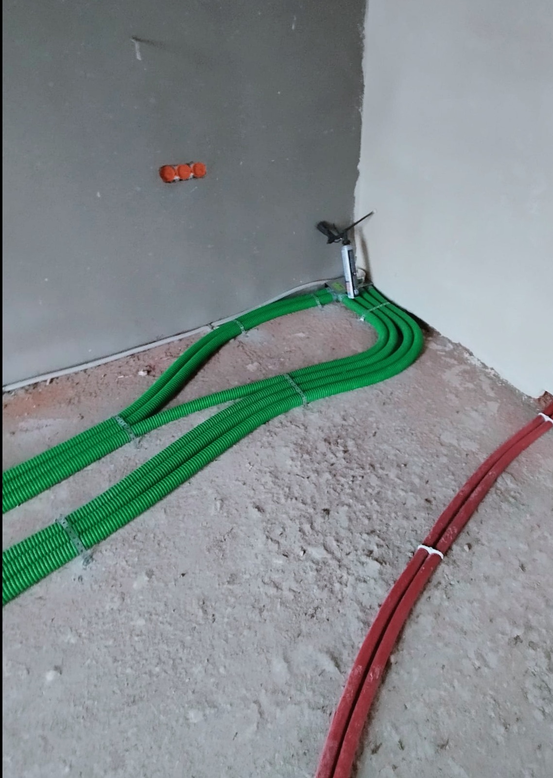 Podłogowe ułożenie zielonych rur karbowanych i czerwonych gładkich rur instalacyjnych w surowym stanie, z widoczną puszką elektryczną w rogu pomieszczenia.