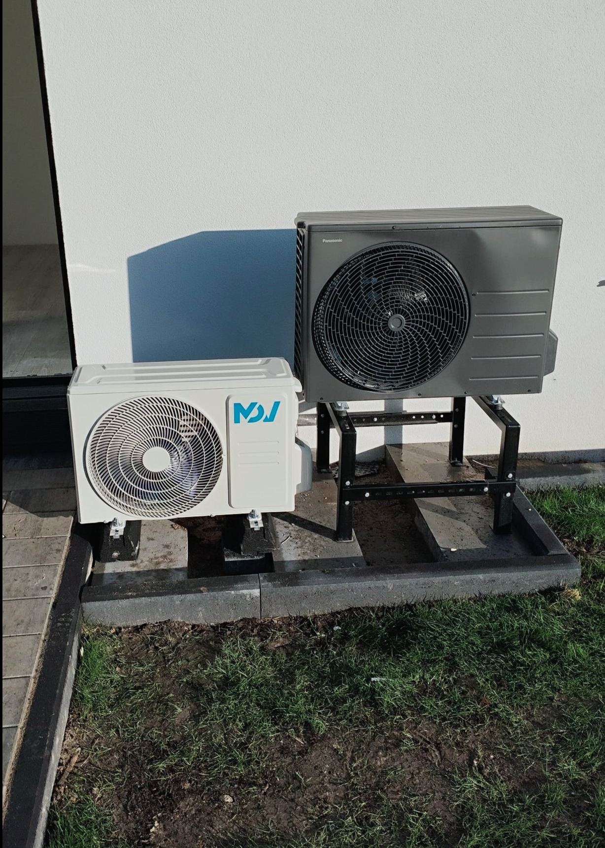 Dwie jednostki zewnętrzne klimatyzacji/wentylacji, jedna biała z niebieskim logo, druga szara Panasonic, zamontowane na metalowych wspornikach na zewnątrz budynku z trawnikiem.