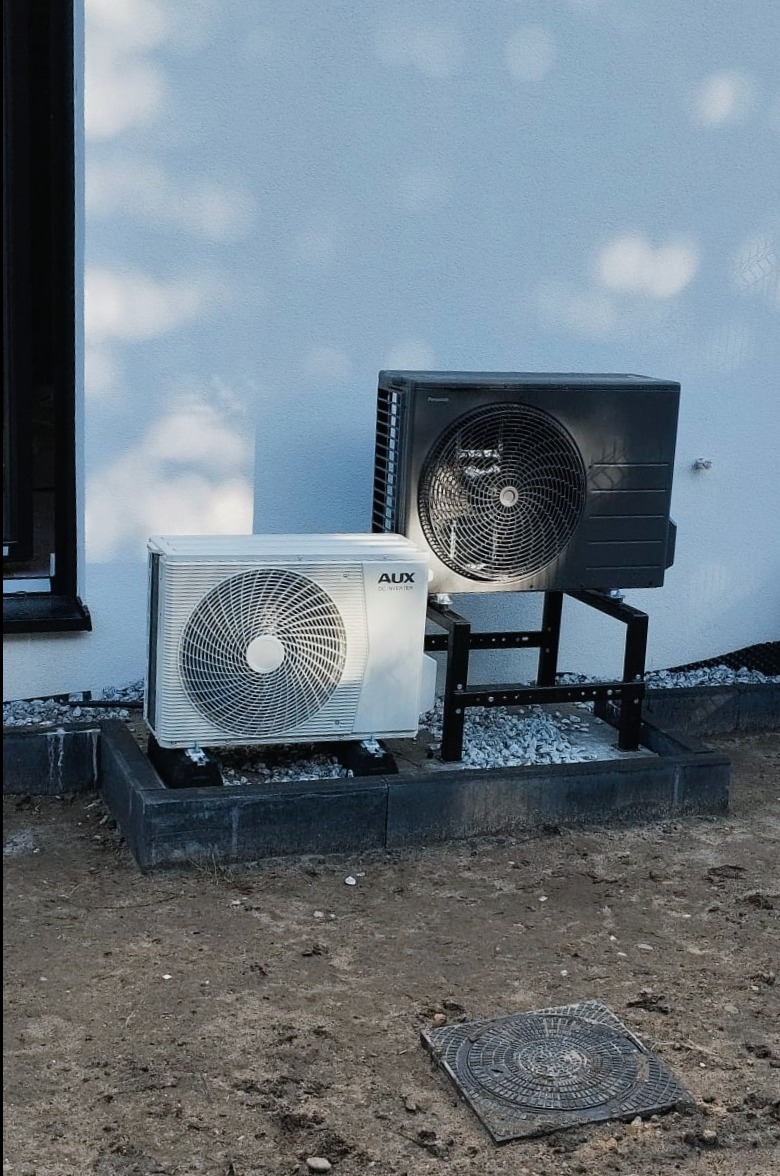Dwie jednostki zewnętrzne pompy ciepła: biała AUX i czarna Panasonic, zamontowane na betonowej podstawie z obramowaniem z kostki i metalowej konstrukcji, na tle białej ściany budynku.