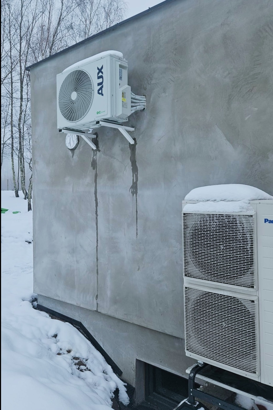 Zewnętrzna jednostka klimatyzacji AUX zamontowana na ścianie budynku z widocznymi śladami kondensatu, obok jednostka Panasonic częściowo przysypana śniegiem, zimowy krajobraz w tle.