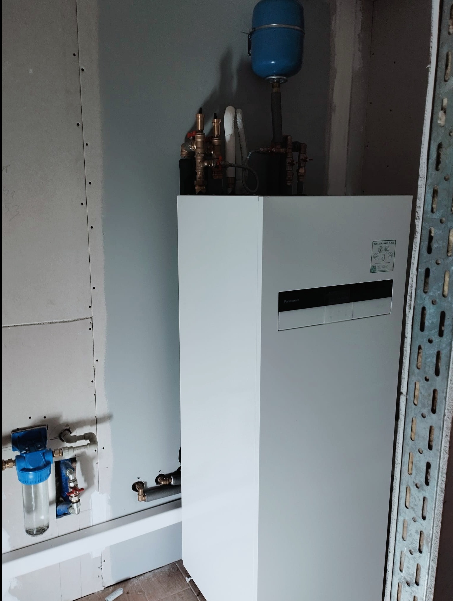 Biała jednostka wewnętrzna pompy ciepła Panasonic Aquarea Smart Cloud, z widocznymi przyłączami hydraulicznymi i zbiornikiem wyrównawczym, zamontowana w pomieszczeniu w trakcie wykańczania.