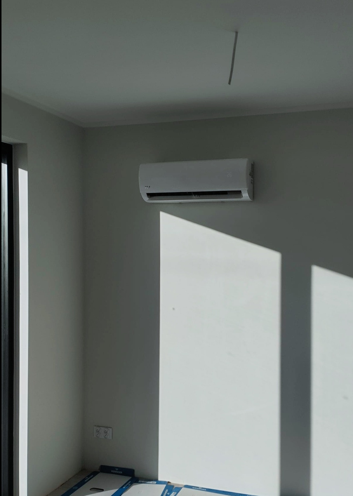 Biała jednostka klimatyzacji zamontowana na jasnej ścianie w nowym pomieszczeniu, widoczne puszki elektryczne i resztki opakowań na podłodze, oświetlona silnym światłem słonecznym wpadającym przez...