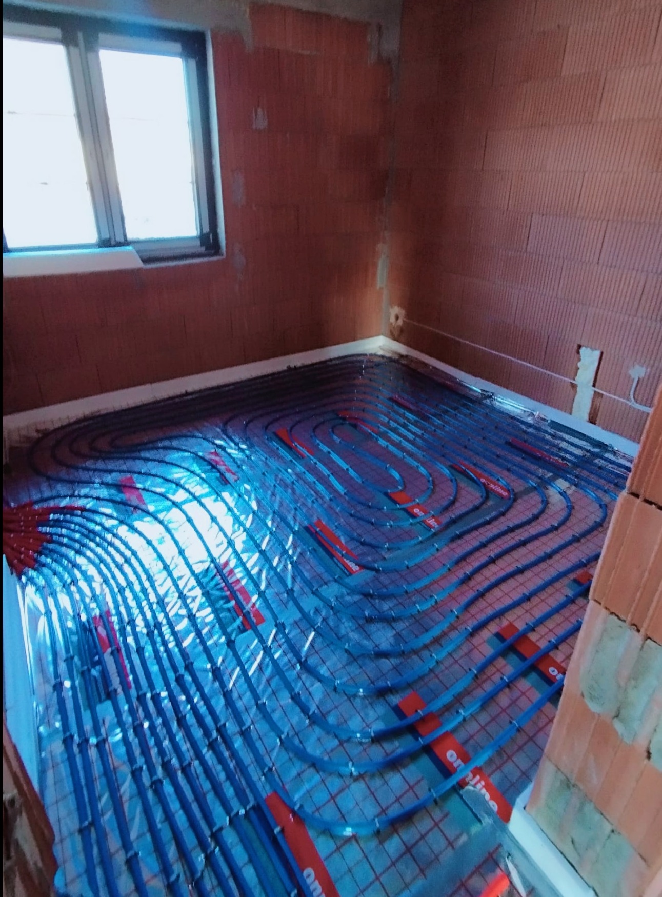 Niedokończona instalacja ogrzewania podłogowego z niebieskimi rurkami i czerwonymi uchwytami na folii odblaskowej w surowym pomieszczeniu z ceglanymi ścianami i oknem.