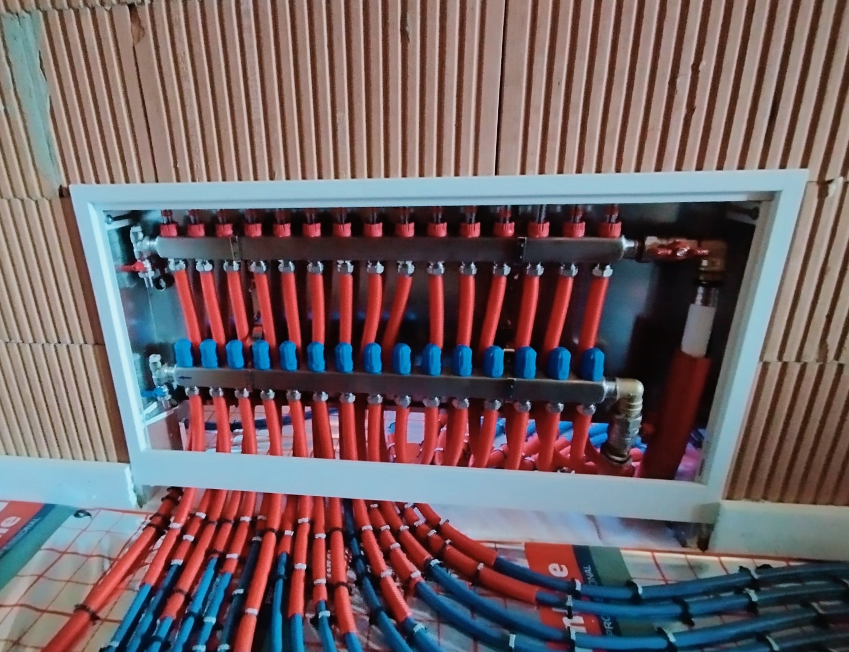 Rozdzielacz ogrzewania podłogowego z czerwonymi i niebieskimi rurami, zamontowany w ścianie z cegły kratówki, widok od dołu.