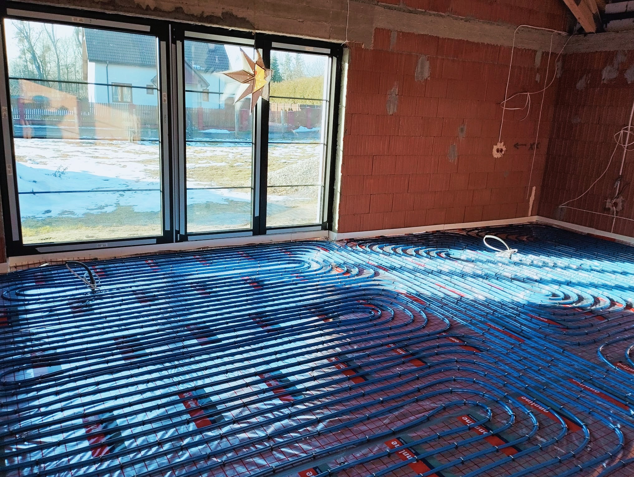 Niebieskie rury ogrzewania podłogowego ułożone na folii w surowym wnętrzu z ceglanymi ścianami i dużymi oknami z widokiem na zaśnieżony ogród.