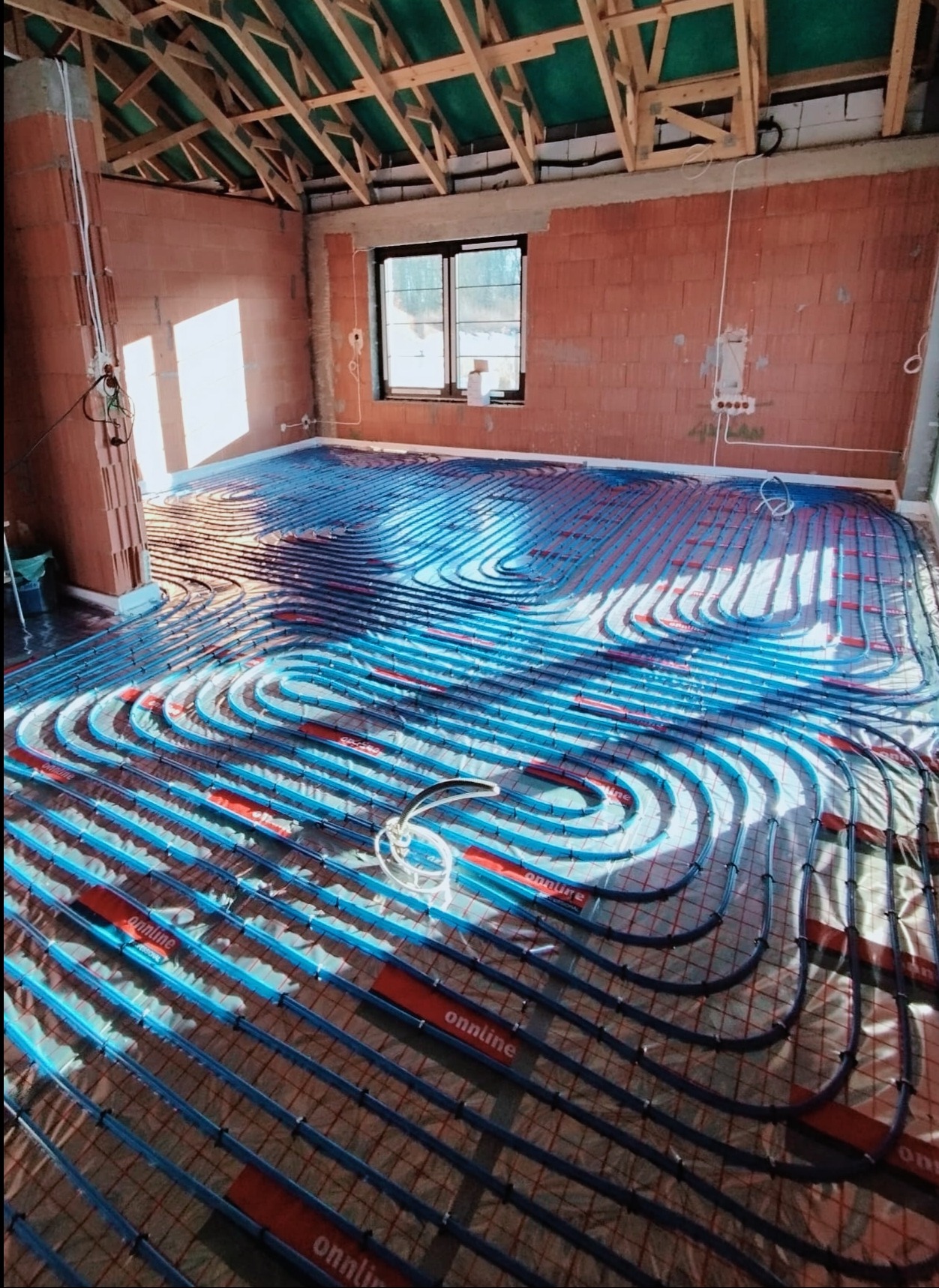 Rozłożona instalacja ogrzewania podłogowego z rurami w kolorze niebieskim i czerwonymi elementami mocującymi, widok na surową cegłę i drewnianą konstrukcję dachu w nowo budowanym domu.