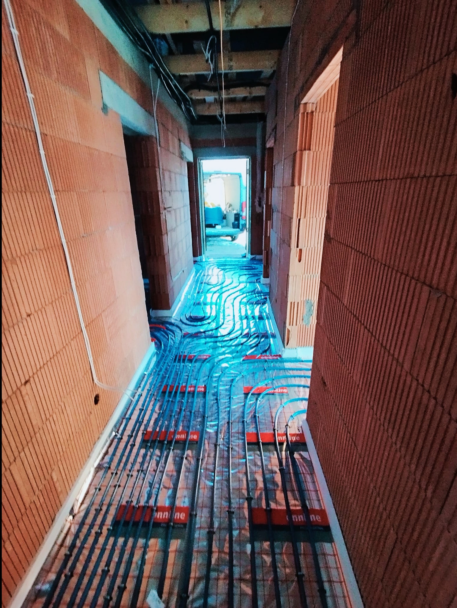 Wnętrze budynku w trakcie budowy z widocznym systemem ogrzewania podłogowego, rury rozprowadzone na folii izolacyjnej, ściany z cegły ceramicznej.