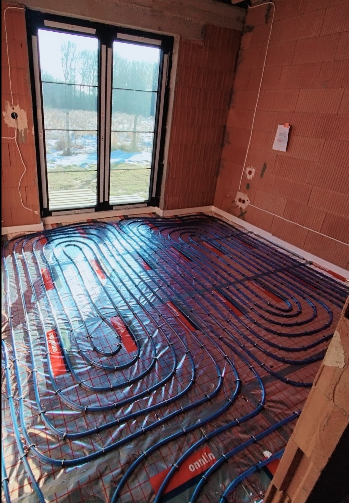 Niedokończona instalacja ogrzewania podłogowego z niebieskimi rurami na folii z czerwoną kratką, widoczne surowe ściany z cegły dziurawki, okno z widokiem na zimowy krajobraz.