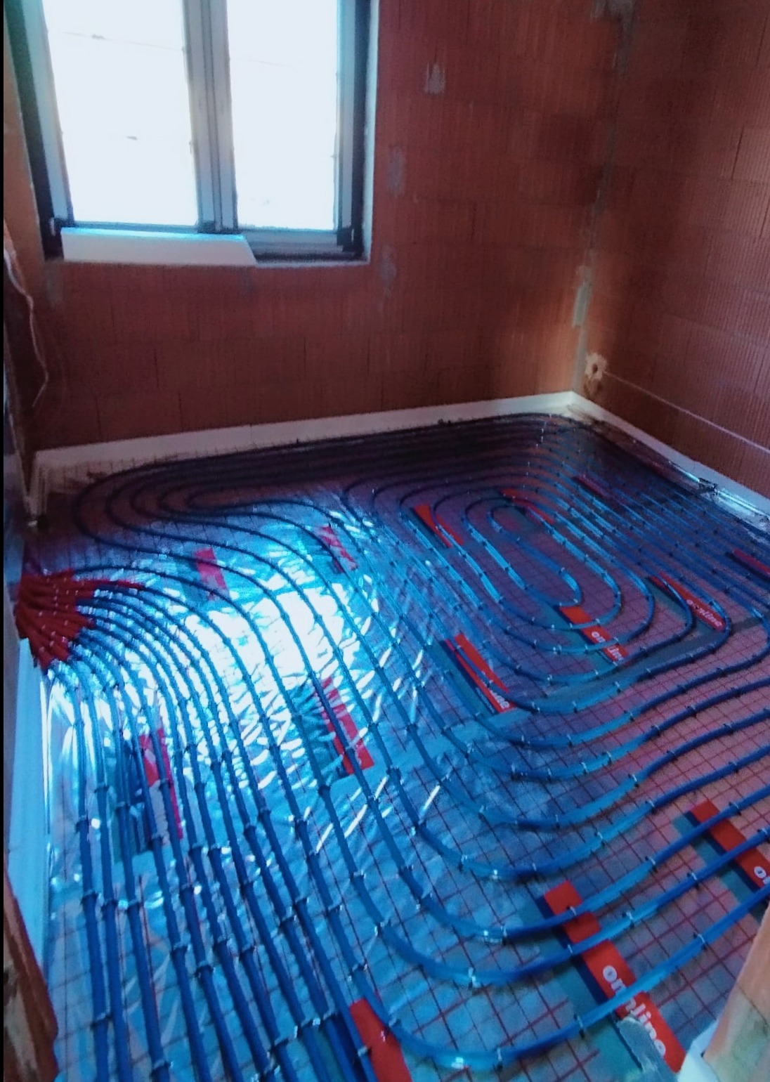 Niebieskie rury ogrzewania podłogowego ułożone na folii izolacyjnej z widocznymi czerwonymi uchwytami mocującymi, na tle surowych ścian z cegły ceramicznej i okna.