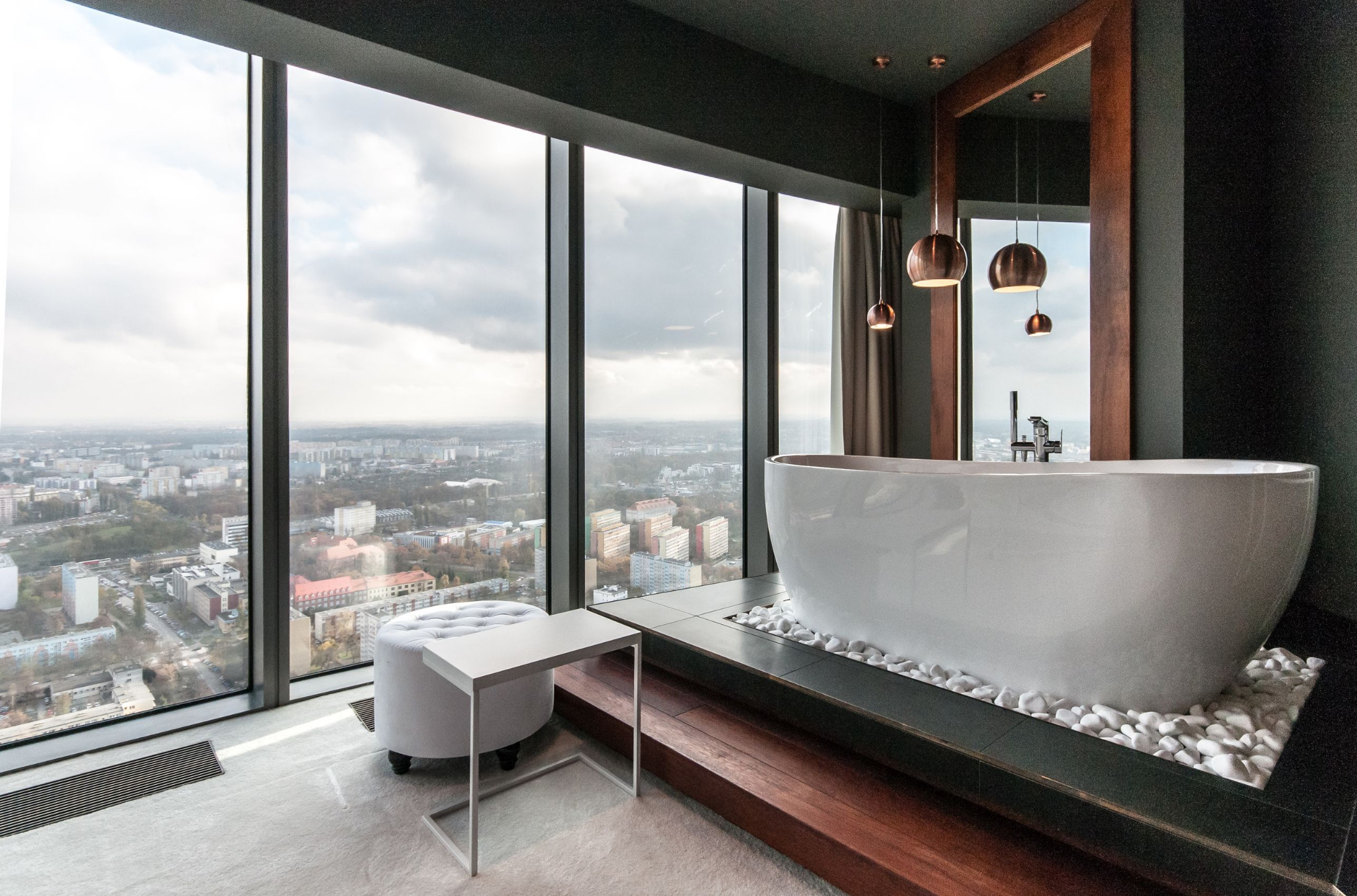 Nowoczesna łazienka w wieżowcu z wolnostojącą wanną na podeście z białych kamieni, widok na panoramę miasta za oknem.