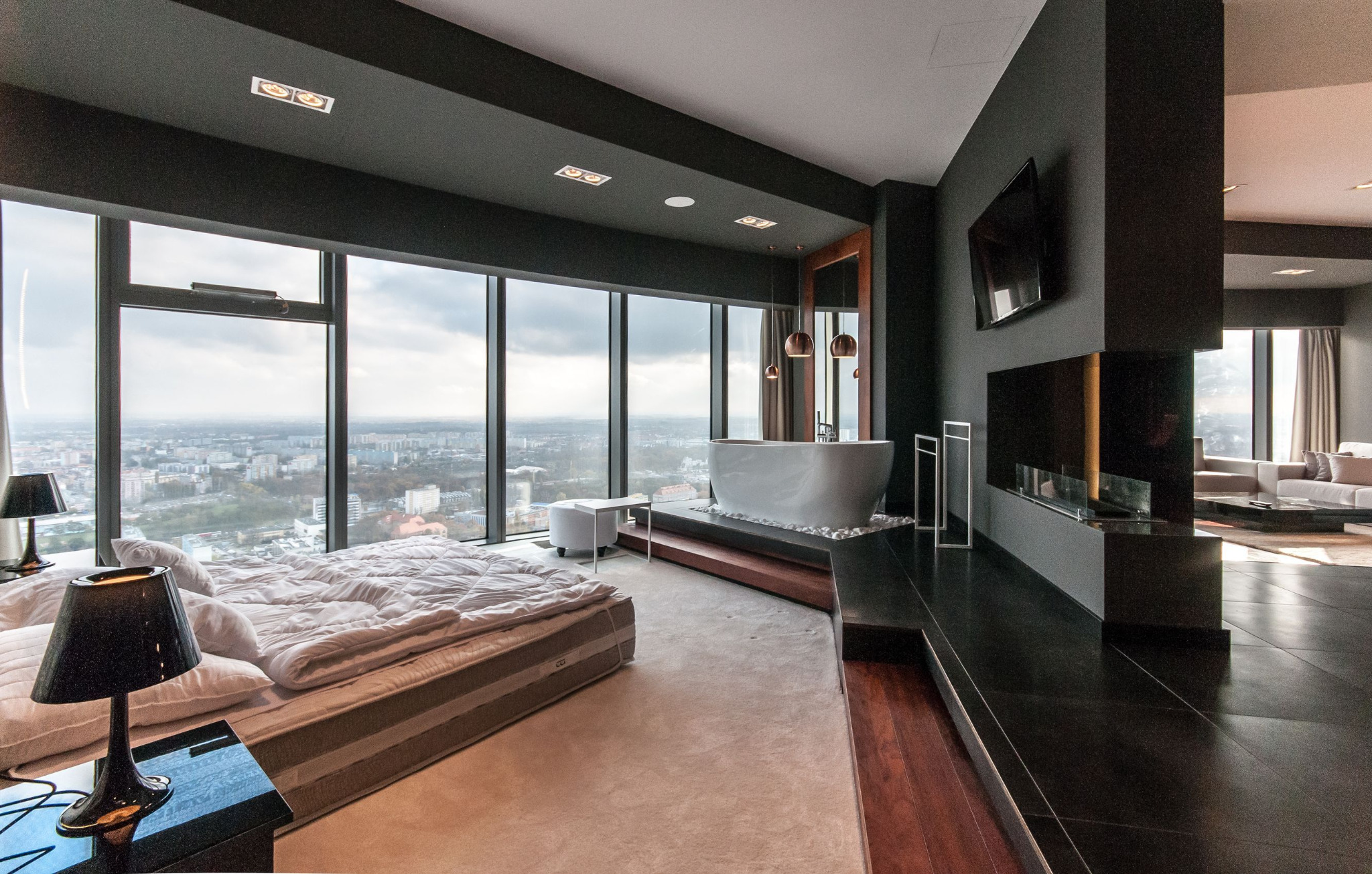 Nowoczesna sypialnia w apartamencie z panoramicznym widokiem na miasto Wrocław, obejmująca łóżko, wolnostojącą wannę na podwyższeniu i kominek.