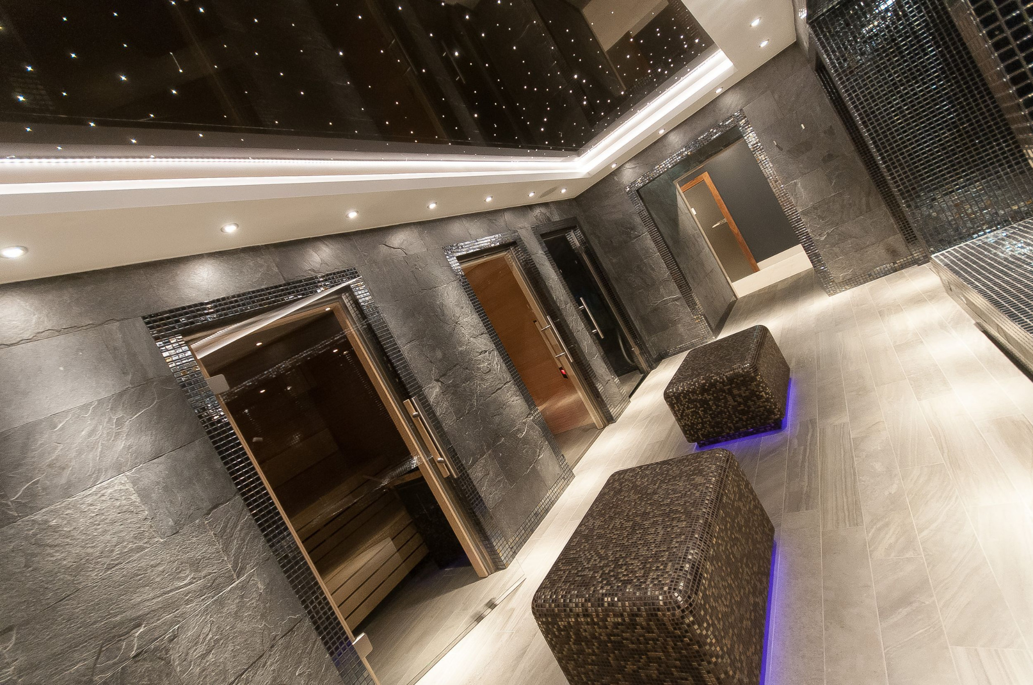 Nowoczesne wnętrze strefy spa z trzema wejściami do saun, ciemnoszare ściany z kamienia łupkowego, mozaikowe obramowania drzwi, sufit z efektem gwieździstego nieba i dwa pufy z mozaiki w brązowych...