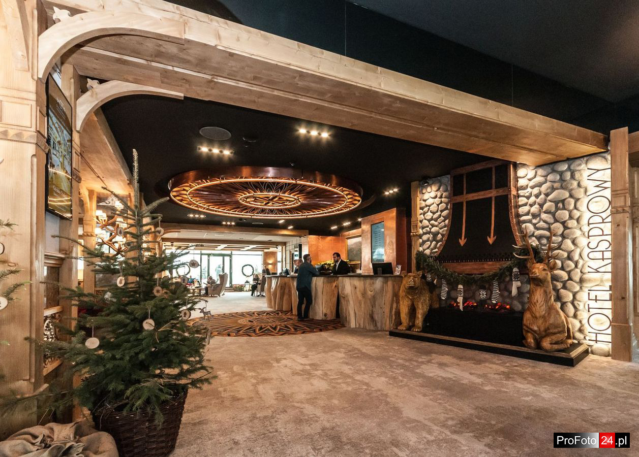 Wnętrze hotelu z recepcją wykończoną drewnem, dekoracyjnym oświetleniem sufitowym i elementami wystroju w stylu góralskim, w tym rzeźby niedźwiedzia i jelenia oraz choinka w rogu