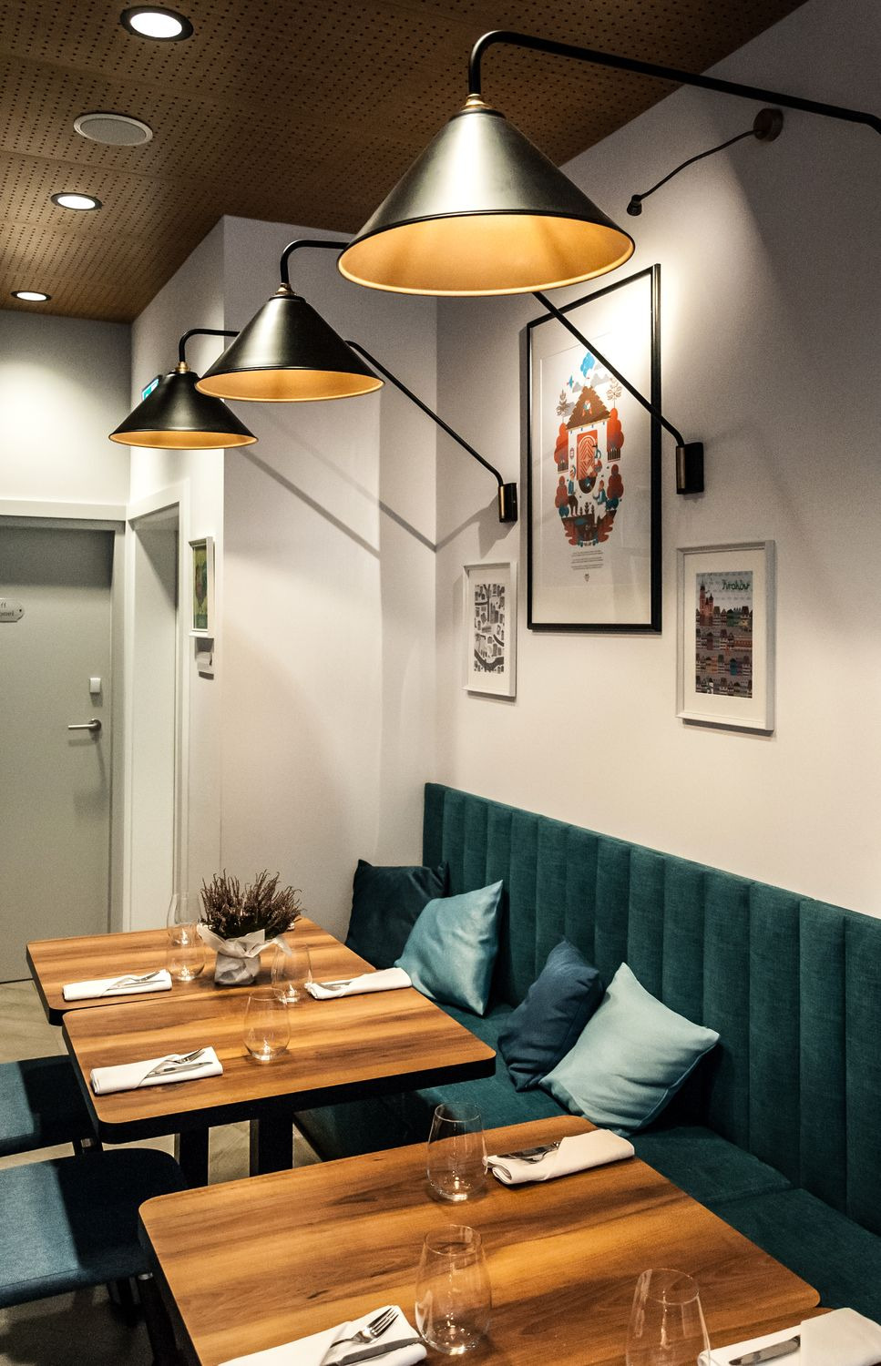 Wnętrze restauracji: drewniane stoły z nakryciami, turkusowa tapicerowana ława z poduszkami, czarne lampy wiszące w kształcie stożków i grafiki na ścianie.