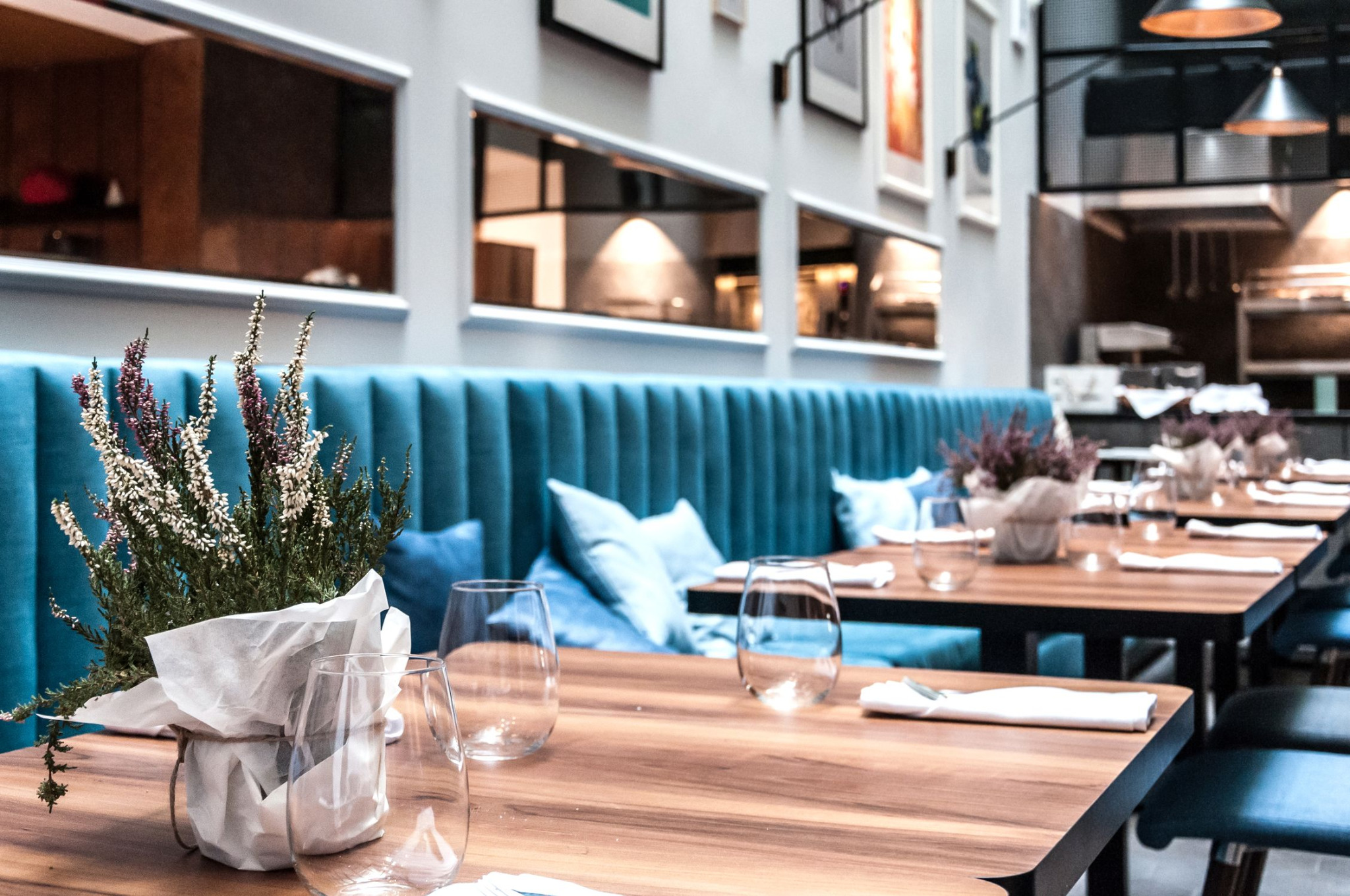 Elegancko nakryty stół w restauracji z turkusową tapicerowaną ławą w tle, dekorowany bukietem wrzosów w białym papierze i szklankami.