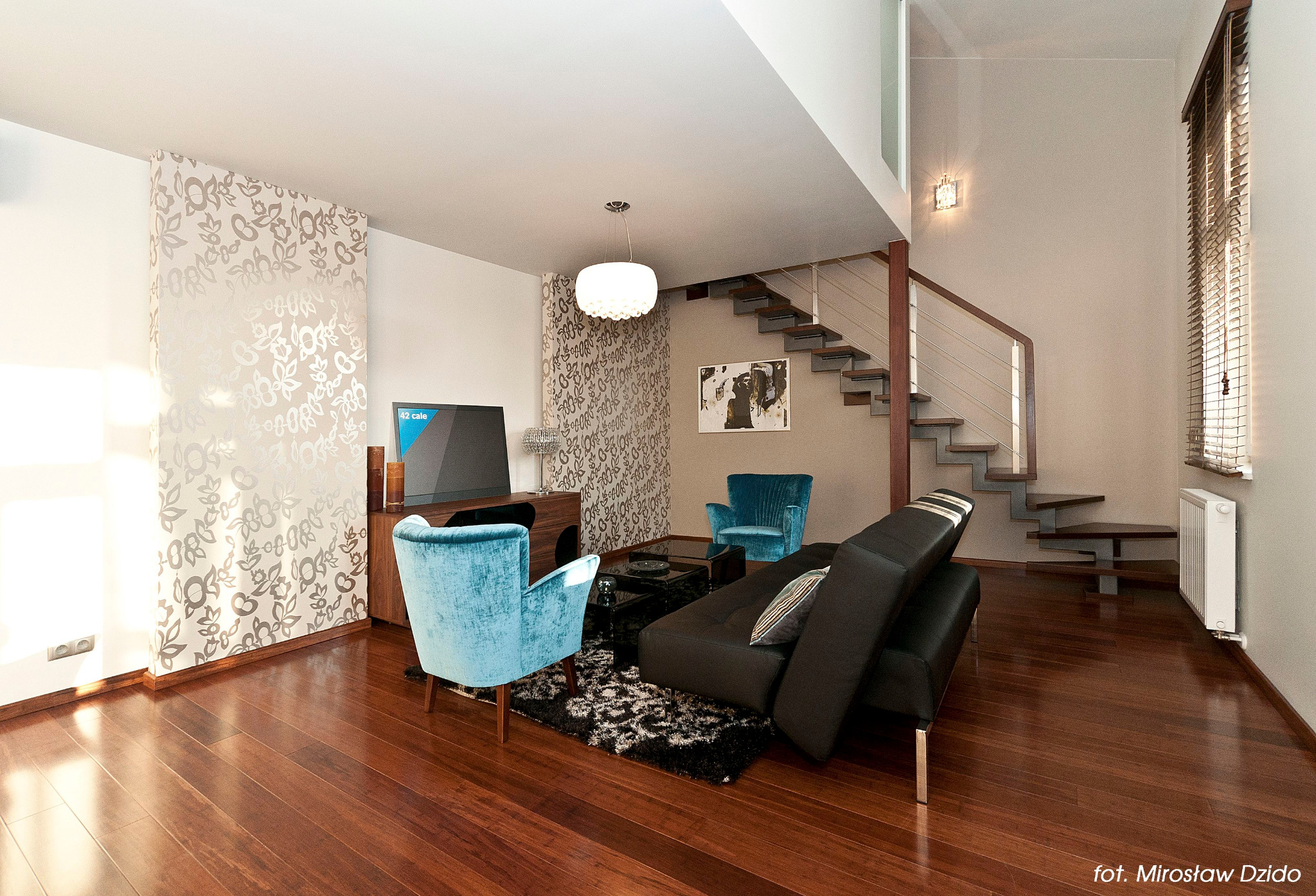 Jasne, dwupoziomowe wnętrze salonu z drewnianą podłogą, turkusowymi fotelami, czarną sofą i nowoczesnymi schodami z metalową balustradą.