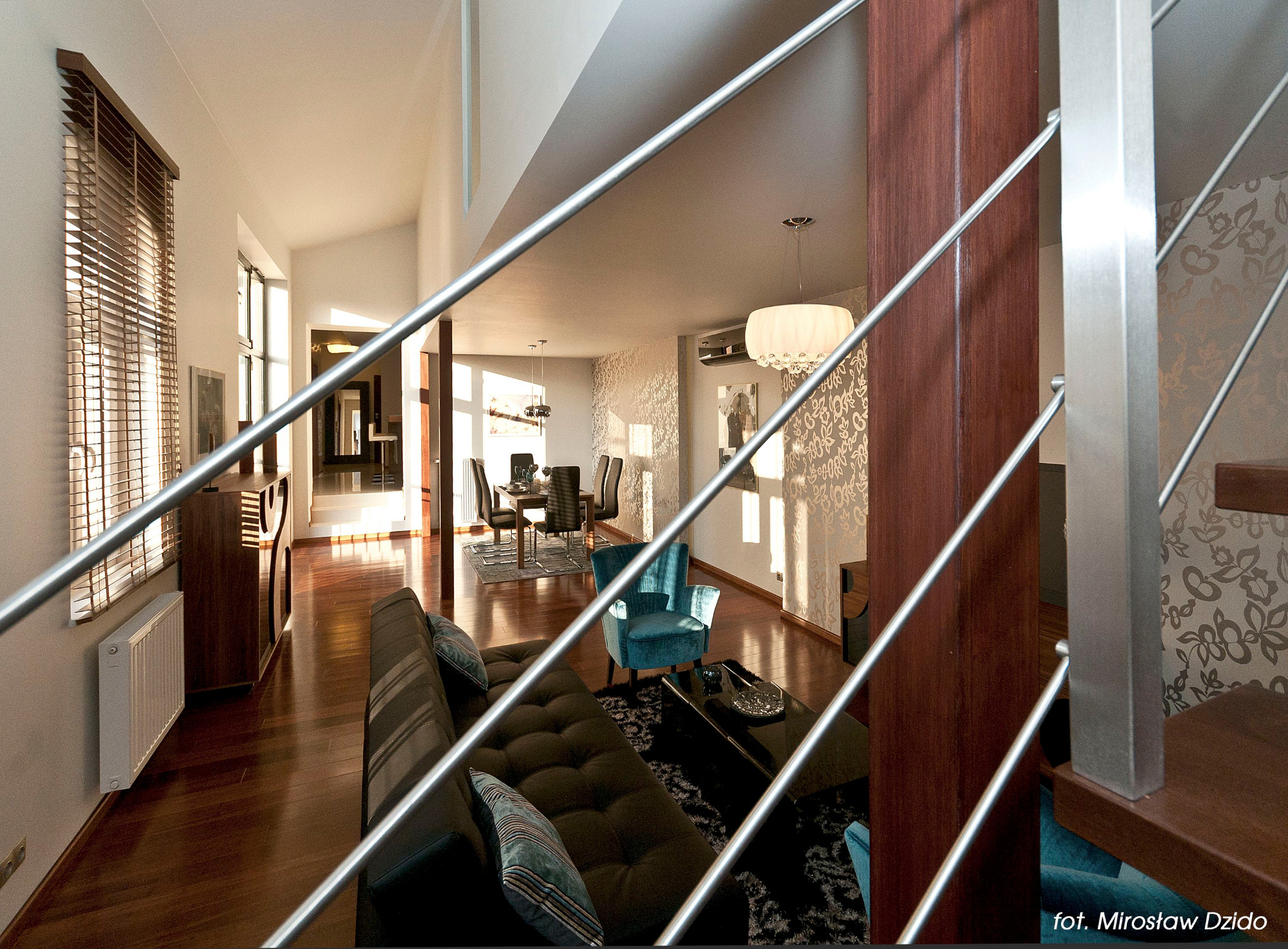 Jasne wnętrze domu z drewnianą podłogą, schodami z metalową balustradą, ciemną sofą, turkusowym fotelem i jadalnią w tle.