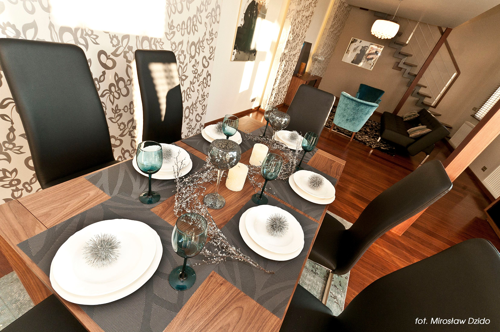 Elegancko nakryty stół z ciemnoszarymi podkładkami, białymi talerzami z srebrnymi dekoracjami, turkusowymi kieliszkami, świecami i srebrną girlandą, w tle salon z schodami i designerskimi fotelami.