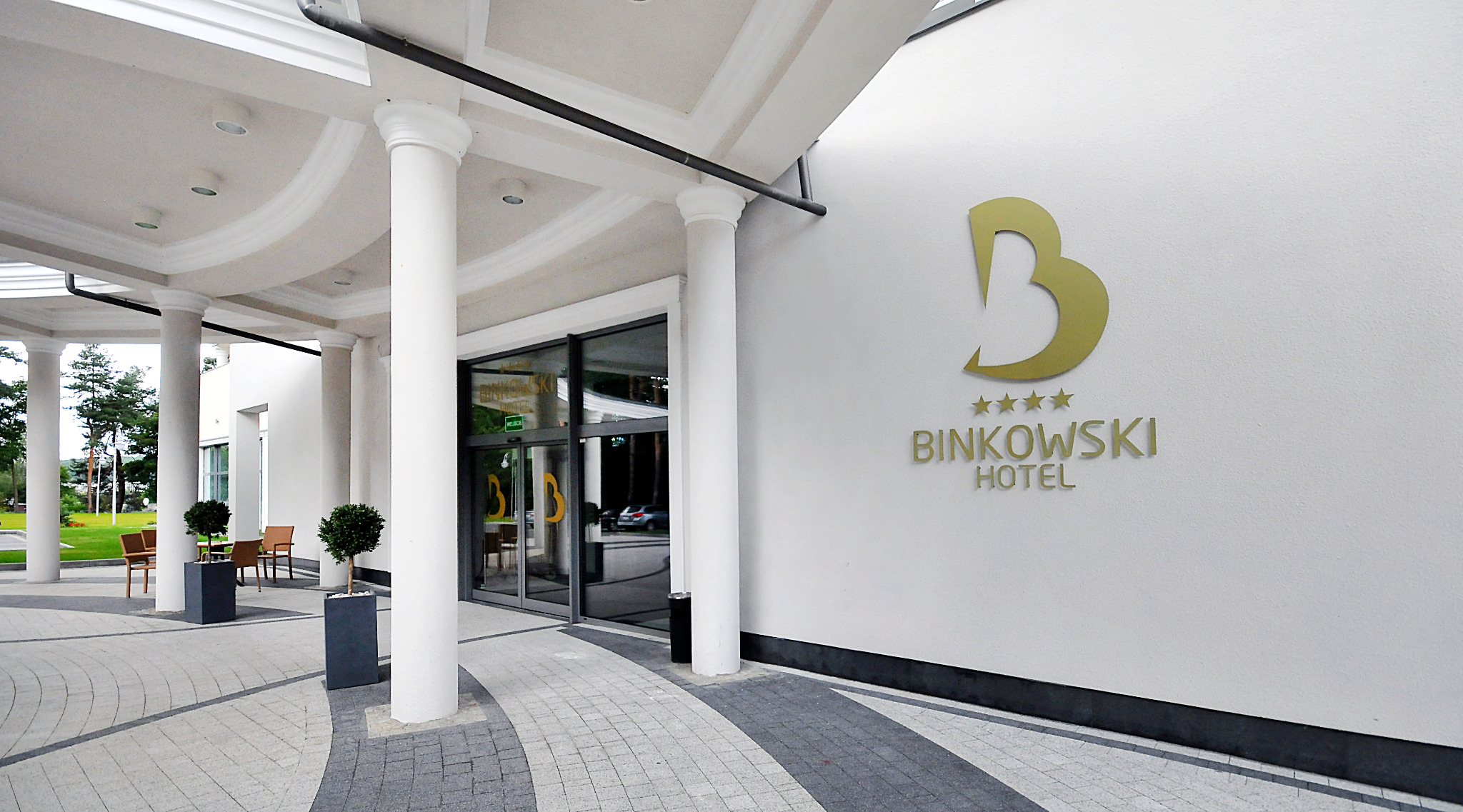 Eleganckie wejście do hotelu Binkowski z kolumnami, roślinami w donicach i złotym logo na białej ścianie.