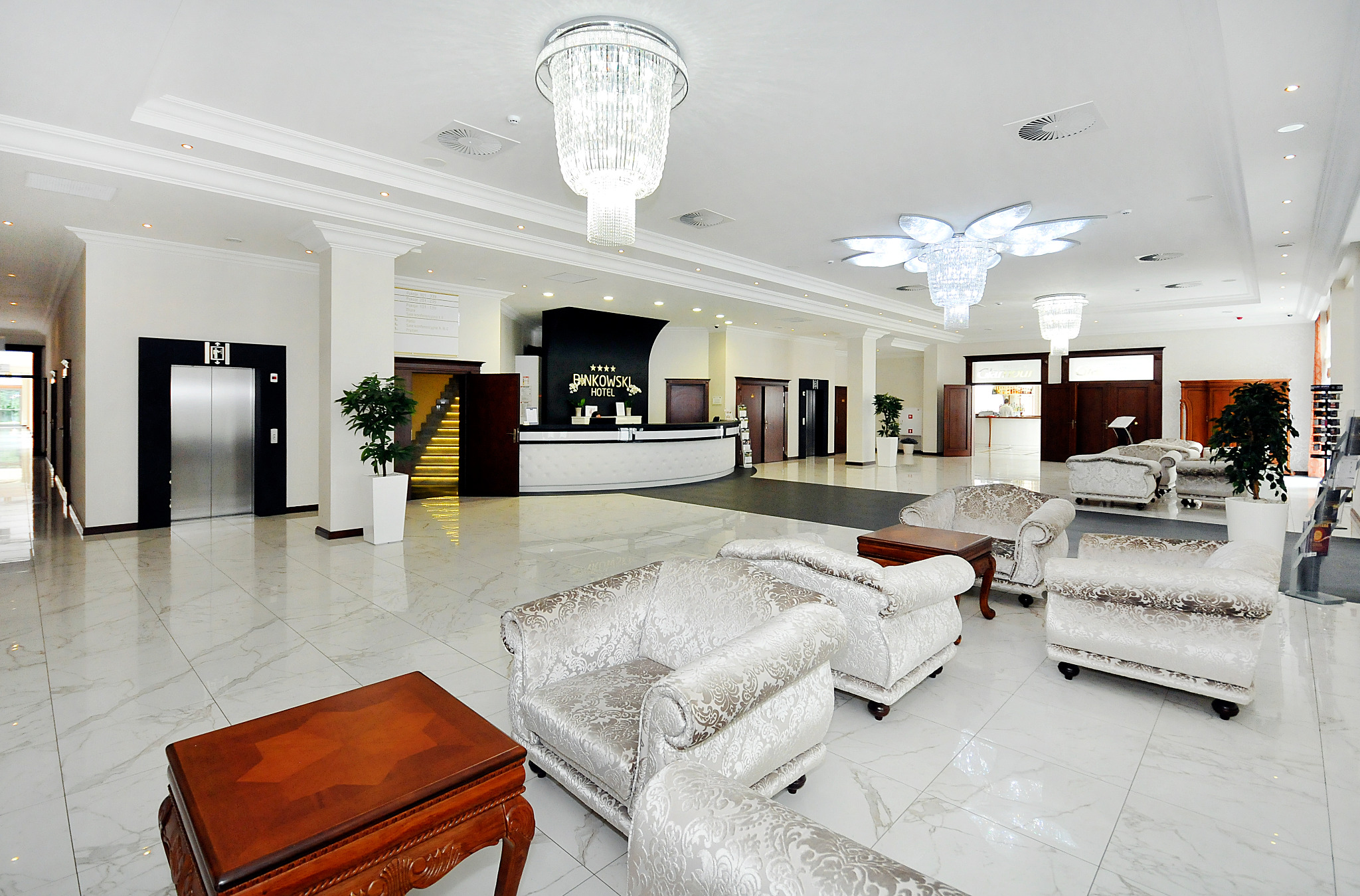 Jasne, przestronne lobby hotelowe z marmurową podłogą, kryształowymi żyrandolami, eleganckimi fotelami w stylu Ludwika i recepcją z logo hotelu Bankowski.