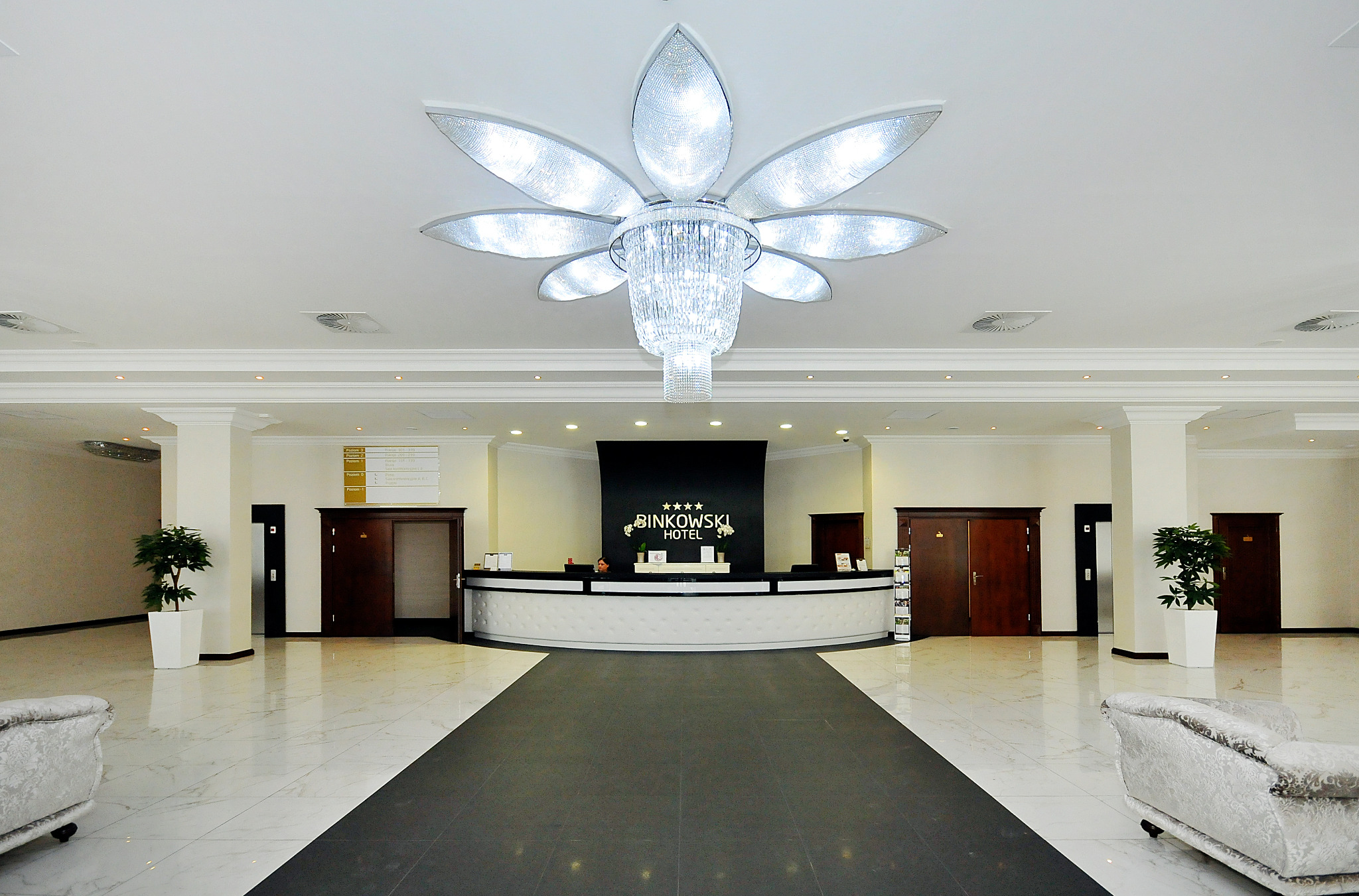 Eleganckie lobby hotelowe z marmurową podłogą, kryształowym żyrandolem w kształcie kwiatu i recepcją z logo Binkowski Hotel.