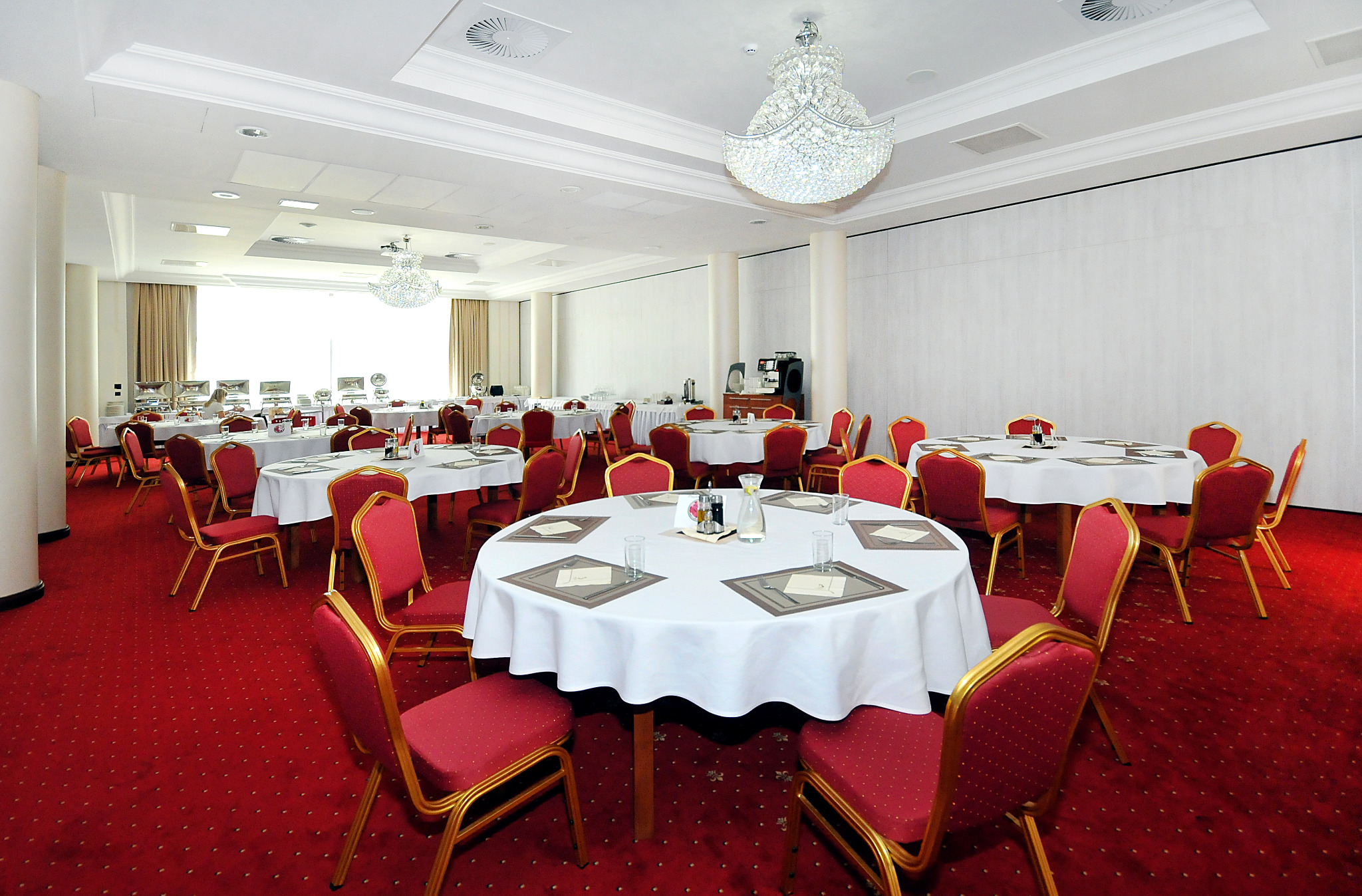 Sala bankietowa z okrągłymi stołami nakrytymi białymi obrusami, czerwone krzesła ze złotymi ramami, żyrandole kryształowe, czerwona wykładzina z wzorem.