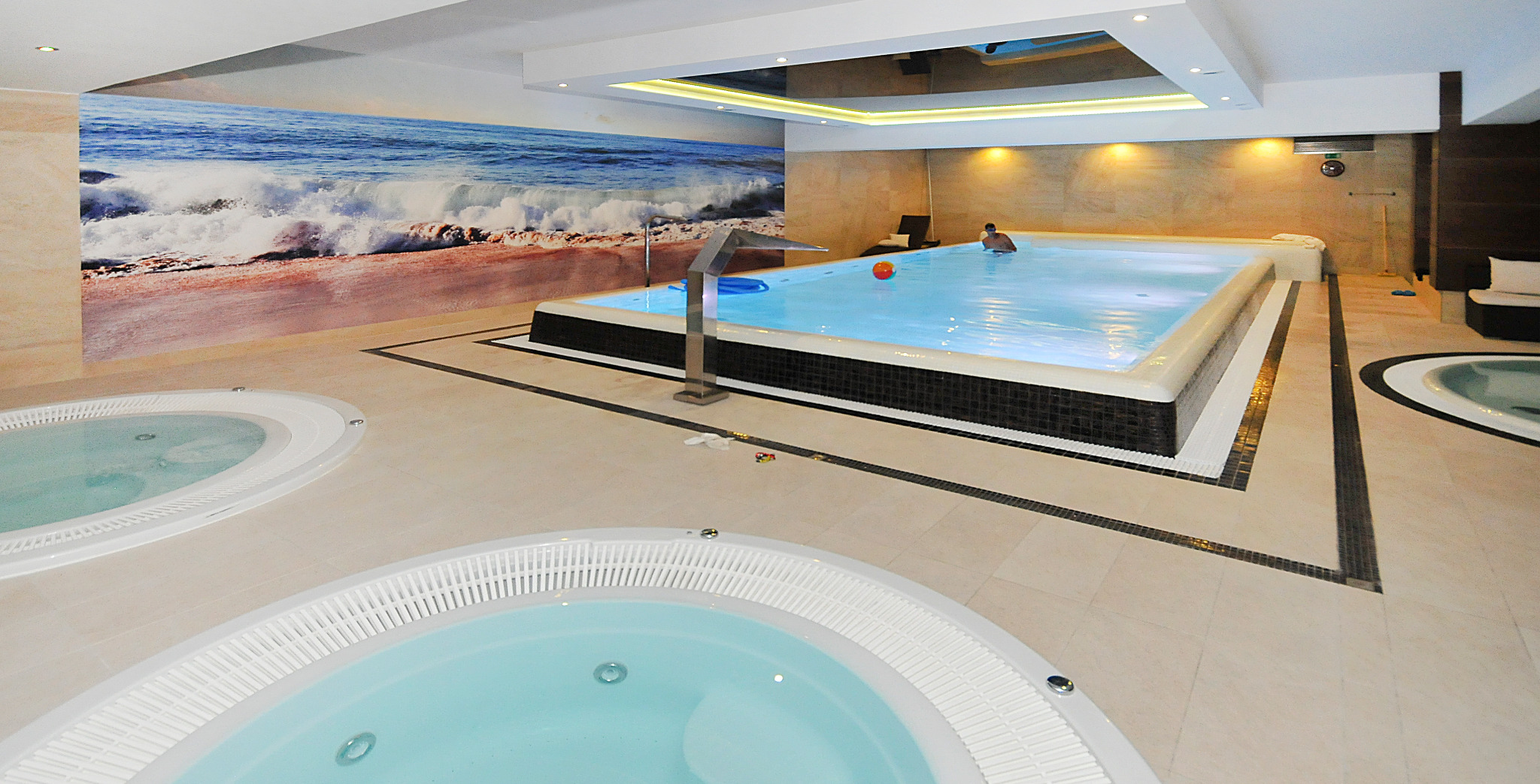 Wnętrze luksusowego spa z basenem i dwoma jacuzzi, ściana ozdobiona fototapetą z motywem plaży i fal morskich, nowoczesne oświetlenie sufitowe.