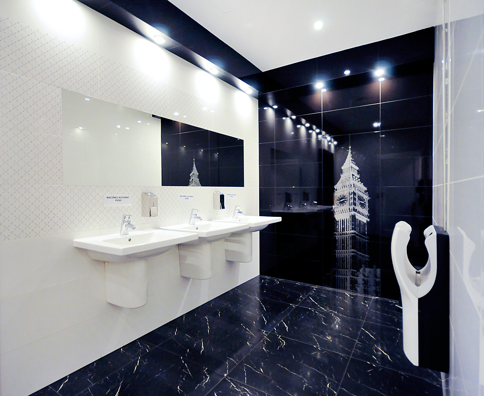 Nowoczesna łazienka publiczna z trzema białymi umywalkami, czarną marmurową podłogą i ścianą z motywem Big Bena w Londynie.