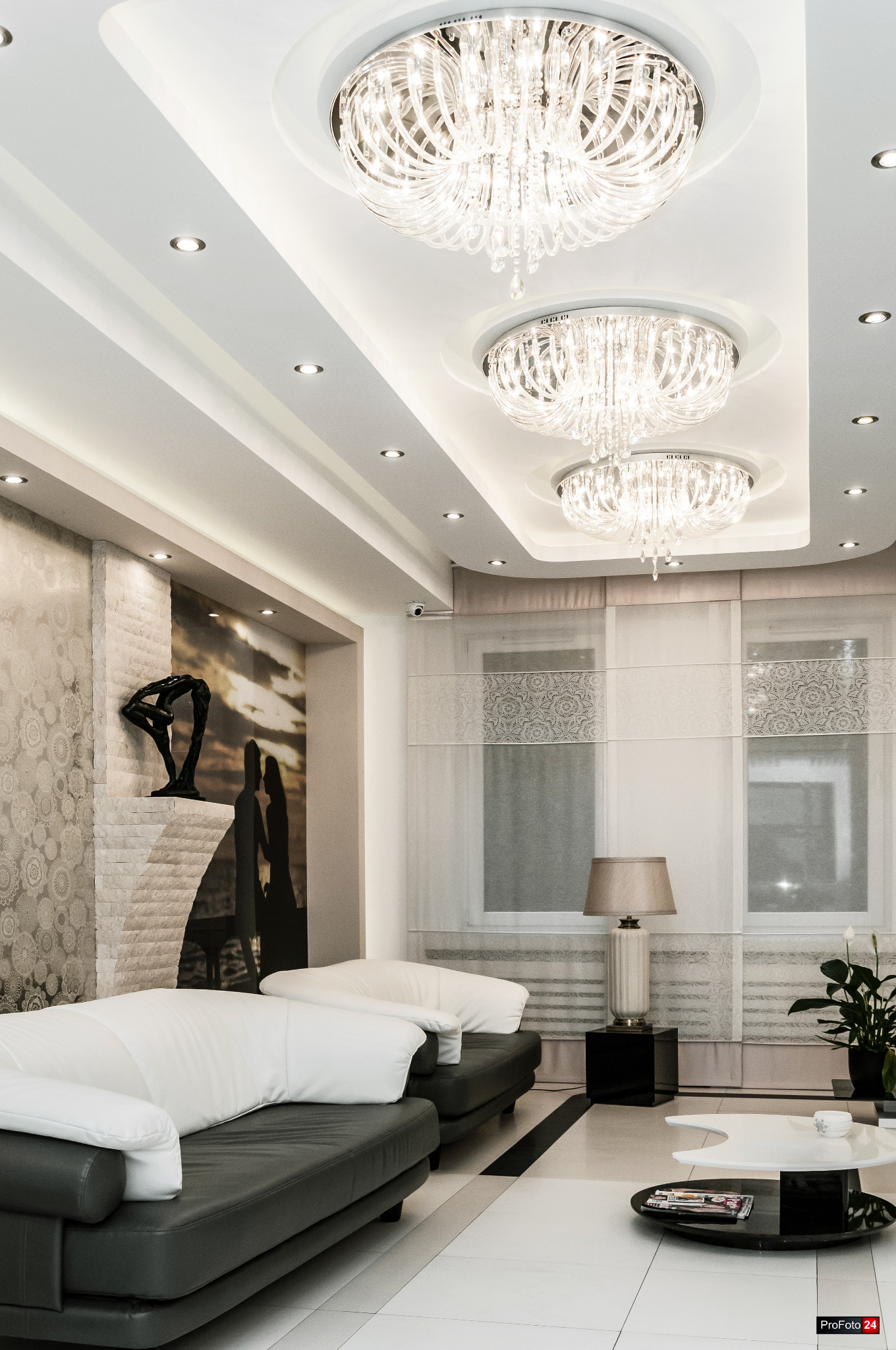 Eleganckie wnętrze z trzema kryształowymi żyrandolami, dwoma skórzanymi sofami z białymi poduszkami, dekoracyjnym kominkiem ze statuetką i abstrakcyjnym obrazem na ścianie oraz stolikiem kawowym...