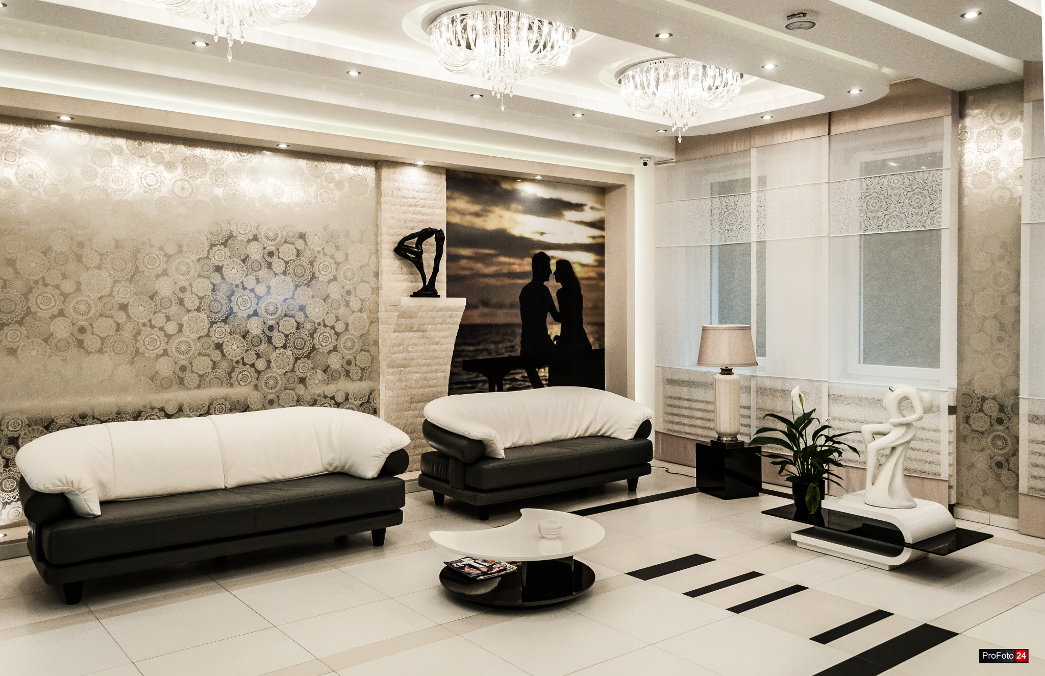 Eleganckie wnętrze salonu z dwoma czarnymi sofami z białymi poduszkami, okrągłym stolikiem kawowym, ozdobną ścianą z motywem kwiatowym, abstrakcyjną rzeźbą i fotografią pary na tle zachodu słońca.