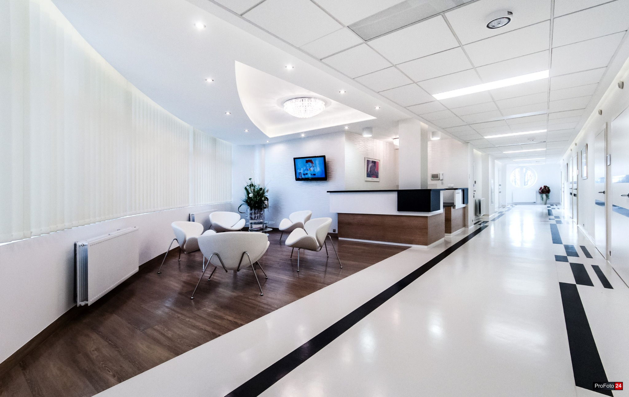 Jasne wnętrze kliniki z białymi krzesłami, recepcją i dekoracyjnym oświetleniem, widok na korytarz z roślinami w tle.