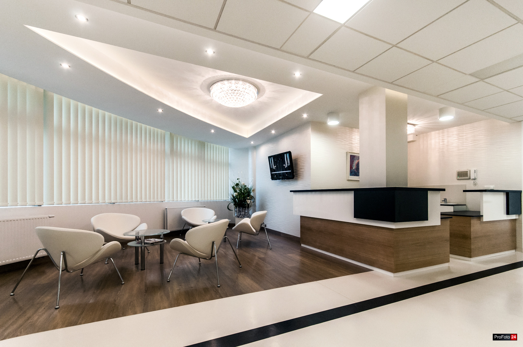 Jasne, nowoczesne wnętrze poczekalni z białymi krzesłami, szklanym stolikiem, recepcją z drewnianym panelem i dekoracyjnym oświetleniem sufitowym.