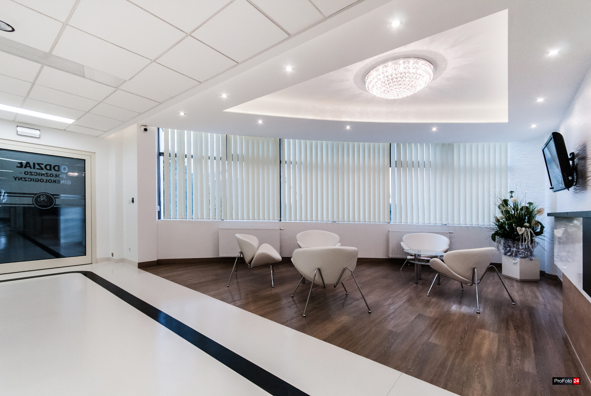 Jasne, nowocześnie urządzone wnętrze poczekalni z białymi krzesłami o unikalnym kształcie, drewnianą podłogą i dużym oknem z żaluzjami w tle.