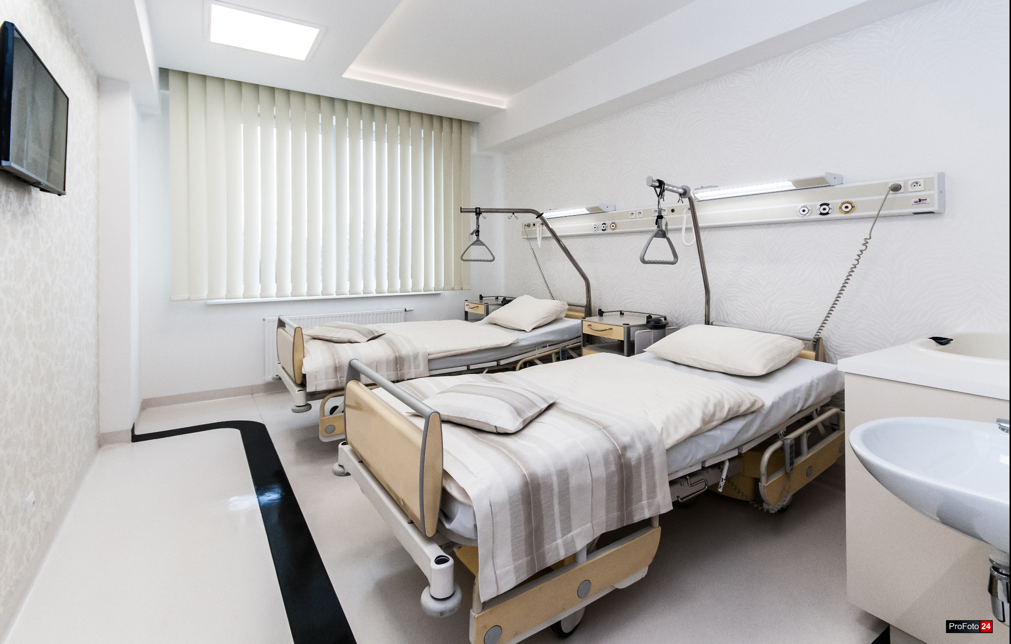Jasne, przestronne wnętrze pokoju szpitalnego z dwoma łóżkami, stolikami nocnymi i umywalką; białe ściany z subtelnym wzorem, rolety w oknie i telewizor na ścianie.