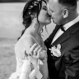 Sara Byczkowska Fotografia - Czarno-białe zbliżenie całującej się pary młodej: panna młoda z ozdobą we włosach i kolczykami w kształcie kwiatów trzyma buty, pan młody w garniturze z butonierką z białej róży.