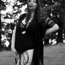 Sara Byczkowska Fotografia - Czarno-białe zdjęcie kobiety w bieliźnie i koronkowym narzutce z frędzlami, stojącej na łące z drzewami w tle, włosy rozwiane przez wiatr.