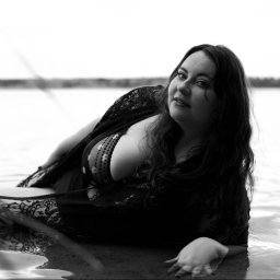 Sara Byczkowska Fotografia - Czarno-biała fotografia kobiety w wodzie, ubrana w koronkowy szal, leżącej na brzegu z fabryką w tle, patrzącej w obiektyw.