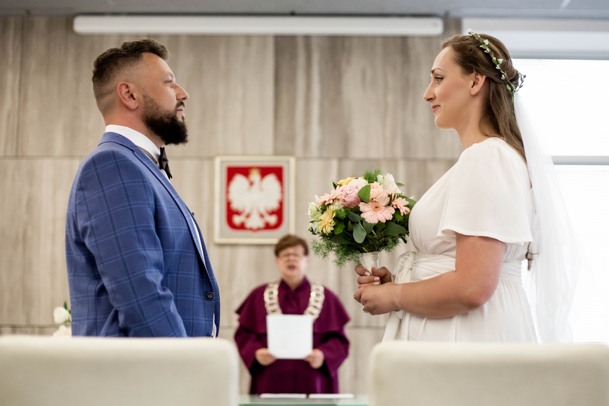 Para młoda podczas ceremonii ślubnej w urzędzie, panna młoda z bukietem kwiatów, pan młody w niebieskiej marynarce w kratę, urzędnik w tle.