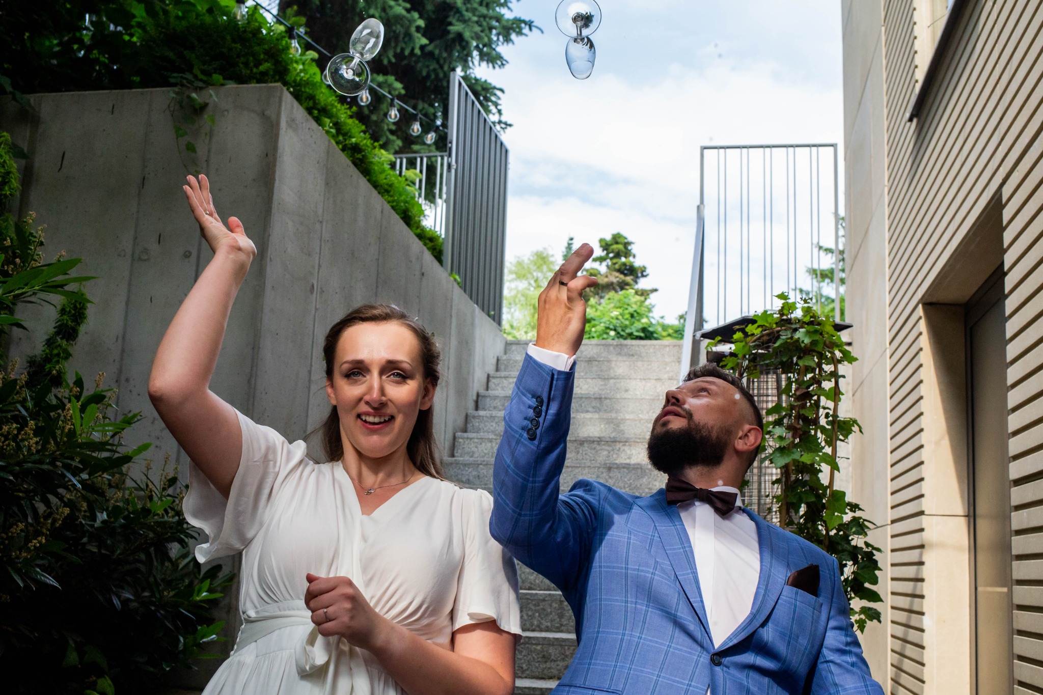 Para młoda, ona w białej sukni, on w niebieskim garniturze w kratę i z muszką, wznoszą ręce do góry, próbując złapać lecące kieliszki do wina na tle betonowej ściany i schodów.