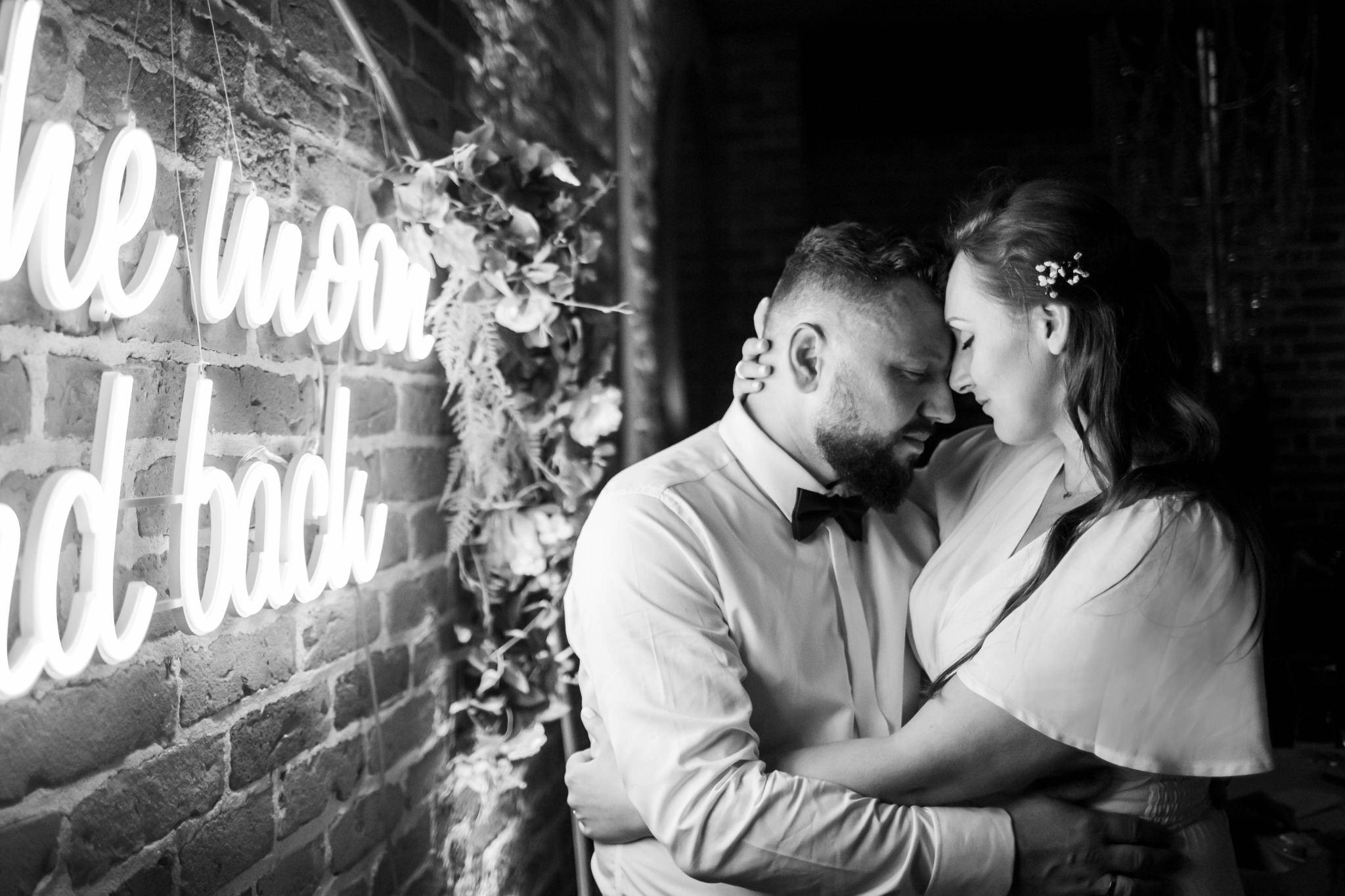 Czarno-białe zdjęcie obejmującej się pary młodej na tle ceglanej ściany z neonowym napisem 'to the moon and back' oraz dekoracją z roślin.