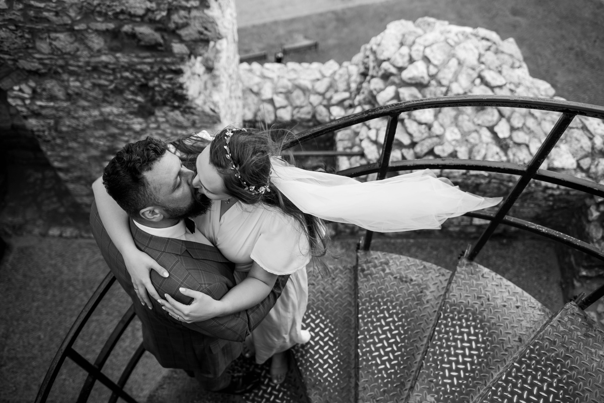 Czarno-białe ujęcie z góry całującej się pary młodej na metalowych schodach spiralnych z kamienną konstrukcją w tle, welon powiewa na wietrze.