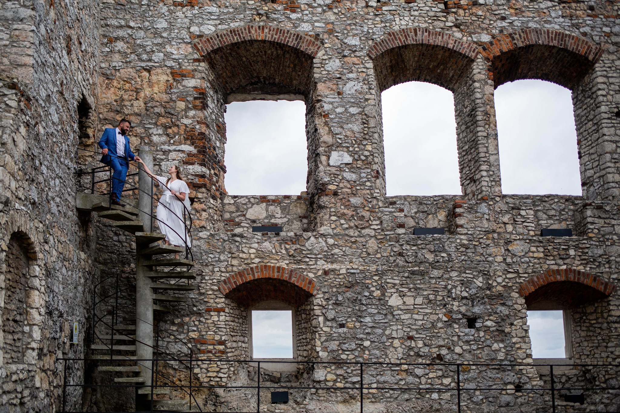 Para młoda na spiralnych schodach w ruinach zamku, on w niebieskim garniturze, ona w białej sukni, trzymają się za ręce, tło z kamiennych murów z oknami.
