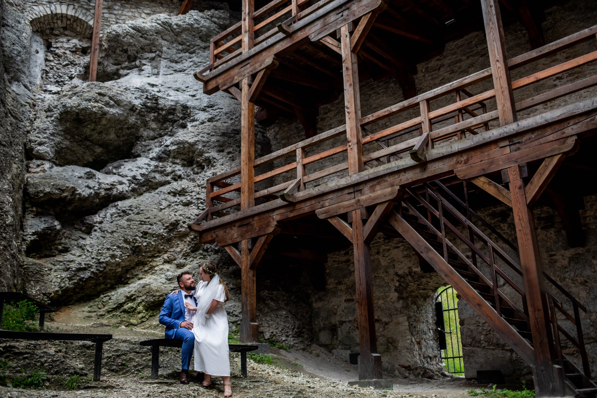 Para młoda siedzi na ławce na tle skał i drewnianej konstrukcji w Koninie, panna młoda w prostej sukni i wianku, pan młody w błękitnym garniturze.