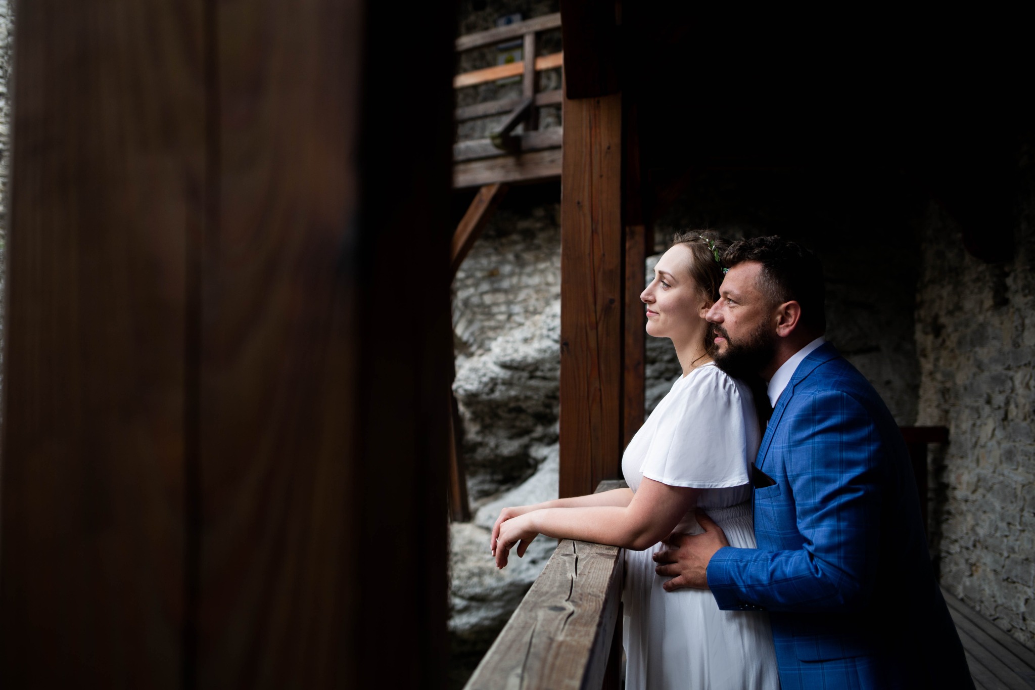 Para młoda na drewnianym balkonie zamku, ona w białej sukni, on w niebieskim garniturze w kratę, oboje patrzą w dal.