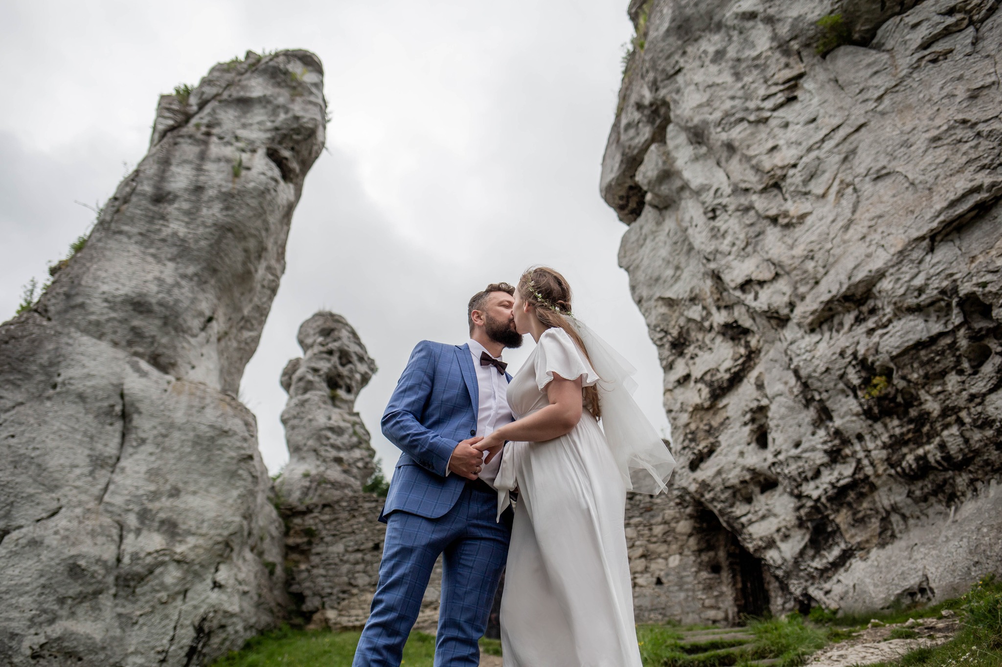 Pocałunek pary młodej w plenerze, on w niebieskim garniturze w kratę i bordowej muszce, ona w białej sukni z welonem, w tle skalne formacje i ruiny zamku.