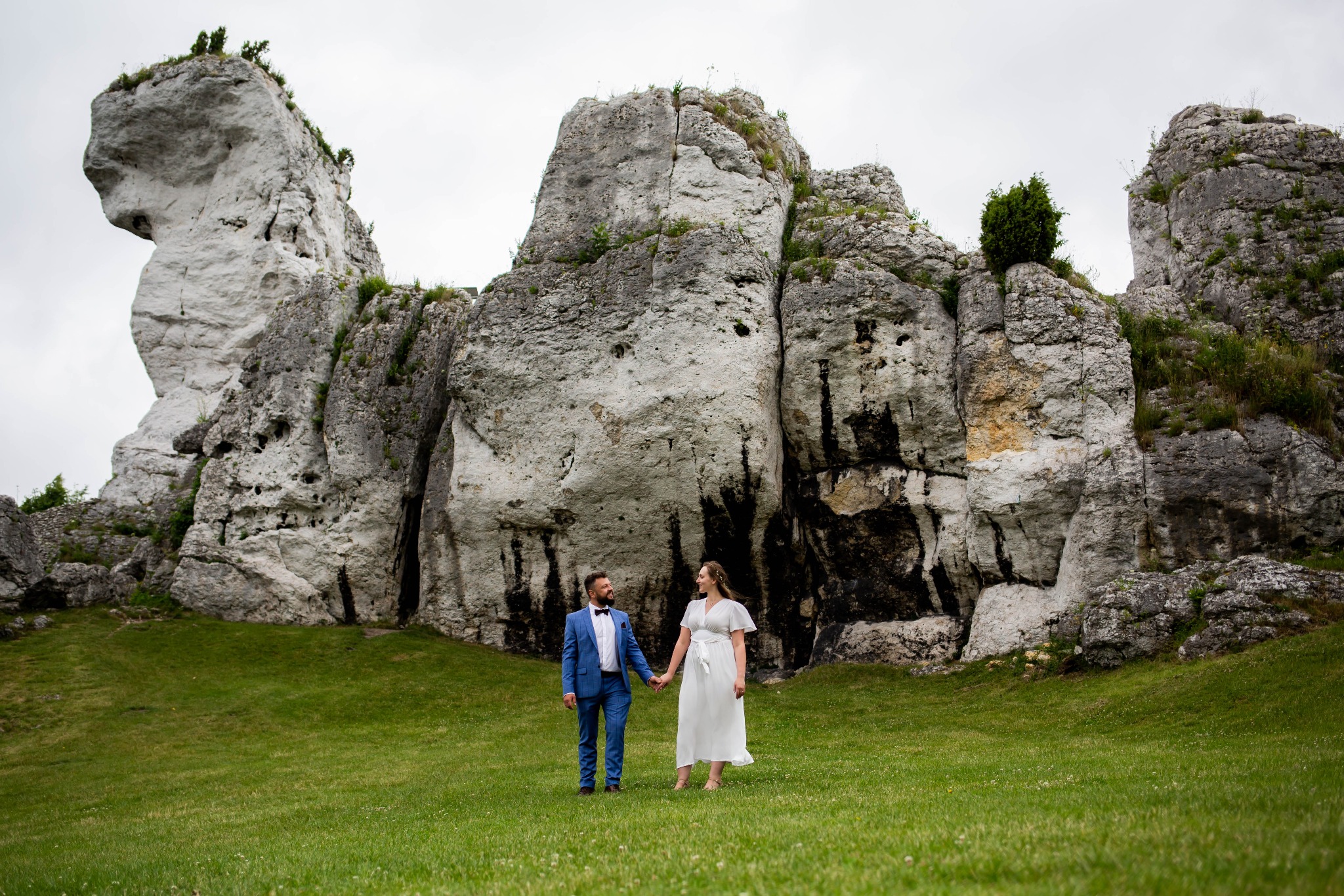 Para młoda trzymająca się za ręce na zielonej łące z wapiennymi skałami w tle, on w niebieskim garniturze i muszce, ona w białej sukience.
