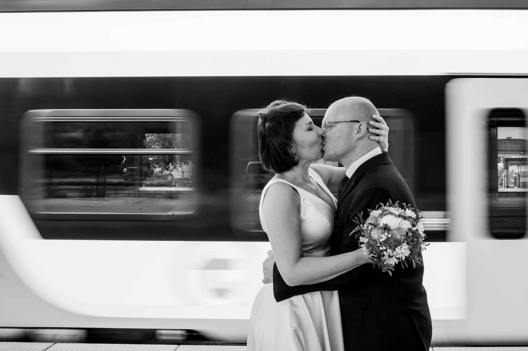 Czarno-białe zdjęcie całującej się pary młodej na tle pędzącego pociągu; panna młoda trzyma bukiet.